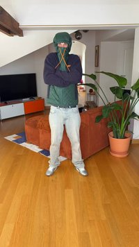 Žalia dygsniuota striukė su gobtuvu, oranžinės spalvos raišteliai, tamsiai mėlynos rankovės, šviesiai mėlyni džinsai ir sidabriniai batai, stovintys patalpose šalia augalo.