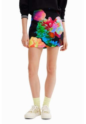 Kvinde iført en sort mininederdel med store farverige blomstermønstre, lysegule sokker, hvide sneakers og en sort langærmet top.