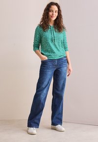 Grüne gemusterte Langarm-Hoodie mit Kordelzügen, kombiniert mit weiten blauen Jeans und weißen Sneakers, vor einem neutralen Hintergrund stehend.
