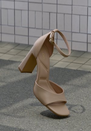 Bézs színű, magas sarkú szandál bokapánttal és vastag sarokkal, csempézett fal előtt, texturált talajon megjelenítve, ferdén elhelyezve.