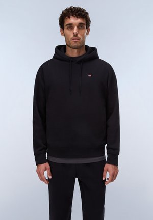 Man with curly hair wearing a black hooded sweatshirt and black pants standing against a light gray background.