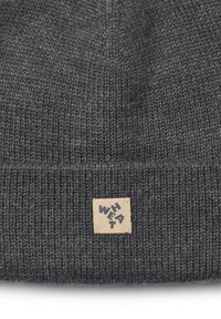 Grijze gebreide beanie met een omgeslagen rand, voorzien van een vierkant tan label met zwarte tekst. De structuur is geribbeld en warm, geschikt voor koude weersomstandigheden.