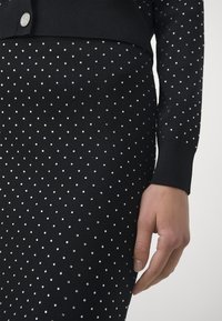 Hand rustend naast een zwart stoffen jurk met lange mouwen, versierd met gelijkmatig verdeelde kleine zilveren studs en een knop met het "MK"-logo.