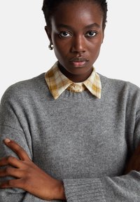 Šedý pletený svetr s žebrovanými manžetami přes žluto-bílou kostkovanou košili. Detailní záběr s překříženýma rukama, zdůrazňující texturu a vrstvený design.