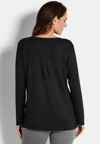 Una donna con capelli lunghi e castani indossa una maglietta nera a maniche lunghe e pantaloni grigi, rivolta di spalle contro uno sfondo grigio chiaro uniforme.