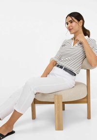Polo à manches courtes rayé en blanc et noir, associé à un pantalon blanc slim-fit et une ceinture marine, assis sur une chaise en bois de couleur claire.