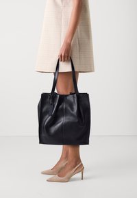 Borsa tote in pelle nera con una finitura testurizzata, due manici lunghi e una chiusura a coulisse, tenuta da una persona che indossa un vestito chiaro.