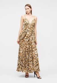 Γυναίκα με μακρύ πλισέ φόρεμα με animal print, λεπτές τιράντες και βαθιά V λαιμόκοψη, φορώντας μαύρα ψηλοτάκουνα σανδάλια, στέκεται μπροστά σε λευκό φόντο.