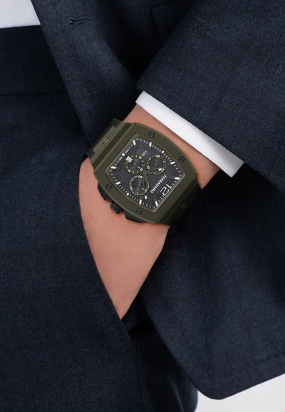 Mees, kes kannab tumedat sinist ülikonda, käsi taskus, näitab rohelist ristkülikukujulist kronograafi käekella musta sihverplaadi ja valgete märgistega.