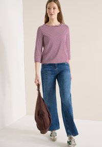 Femme portant un haut à manches longues rayé violet et blanc, un jean bleu, des baskets blanches, tenant un grand sac en daim marron dans une main.