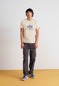 Beige t-shirt med et lilla logo, parret med mørke grå bukser og blå sneakers. Simpelt design, korte ærmer, afslappet pasform.