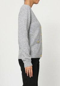 Cardigan en tricot gris avec une coupe ajustée, ornée de boutons dorés et de deux poches plaquées. Il présente des poignets et un ourlet côtelés.