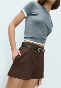 Donna che indossa una maglietta grigia corta e pantaloni corti marroni a vita alta con una cintura nera, con una mano in tasca contro uno sfondo neutro.