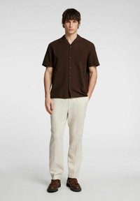 Camicia marrone a maniche corte con chiusura a bottoni, pantaloni beige e scarpe marroni, caratterizzata da una vestibilità comoda e un colletto.