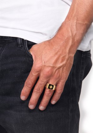 Anneau doré avec une incrustation noire présentant un motif d'étoile éclatante. L'anneau a une finition lisse et polie et une forme rectangulaire.