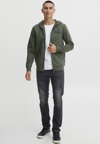 Oliven grøn zip-up hoodie med en frontlomme, grå jeans og sorte sneakers. Hvid indvendig foring og logo patch detalje på brystet.