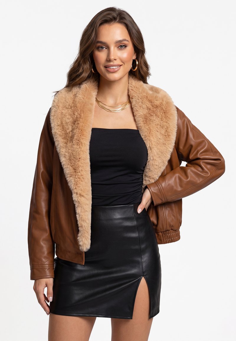 Femme portant une veste en cuir marron avec un col en fourrure beige, un haut bustier noir, une jupe en cuir noire avec une fente sur le devant, ainsi que des boucles d'oreilles créoles et un collier en or.