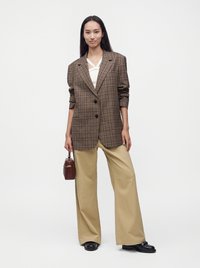 Femme portant un blazer oversize marron à carreaux, un haut blanc, un pantalon beige à jambes larges, des chaussures noires, tenant un petit sac à main marron.