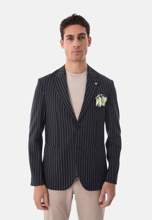 Uomo indossa blazer nero a righe con fazzoletto bianco e giallo nel taschino, sopra camicia beige e pantaloni beige, in piedi di fronte a uno sfondo neutro.