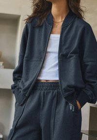 Donkerblauwe zip-up bomberjack met ribgebreide boorden, gecombineerd met bijpassende sweatpants met zijzakken met rits en een witte aansluitende tanktop.