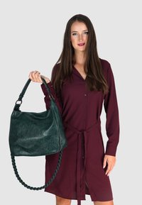 Groene leren handtas met textuur, groot, hobo-vorm, gevlochten handvat. Model draagt een bordeauxrode knoopjurk met een riem.