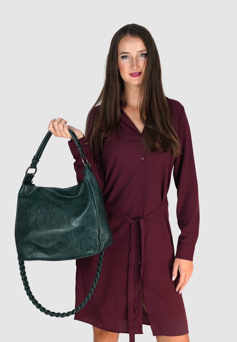Groene leren handtas met textuur, groot, hobo-vorm, gevlochten handvat. Model draagt een bordeauxrode knoopjurk met een riem.