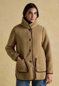 Beeži fliistekk, millel on kõrge krae, pruuni ääristuse, suured esipocketid ja nööpidega kinnitamine. Pehme tekstuur ja mugav lõige.