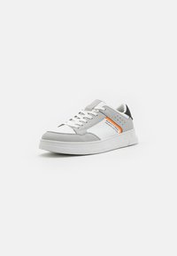 Sneaker grigio e bianco con pelle liscia e accenti in suede. Presenta lacci bianchi, una suola testurizzata e un'etichetta arancione sul lato.