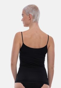 Young person with short blonde hair wearing a black camisole and lace-trimmed black underwear, facing away against a plain background.