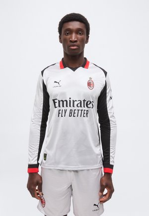 Jugador de fútbol masculino vestido con uniforme blanco de portero del AC Milan con acentos negros y rojos, de pie frente a un fondo claro y sencillo.