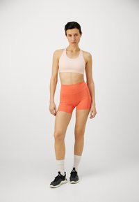 Soutien-gorge de sport en tissu rose clair avec un décolleté arrondi, associé à un short taille haute en orange vif, doté de poches latérales et d'une texture lisse.