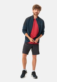 Giacca blu con zip e dettagli neri, t-shirt rossa, pantaloni corti neri e scarpe da trekking blu. Il modello sta in piedi con le mani che tengono la giacca.