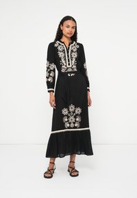 Fekete maxi ruha hosszú ujjal, fehér virágos hímzéssel és testhez simuló derékkal. Lecsúszó alj ráncolt részletekkel. Pántos szandál.