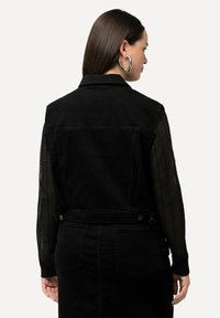 Schwarze Jeansweste mit weitem Kragen, verziert mit transparenten, gestreiften Ärmeln und zwei Gesäßtaschen. Mit silbernen Knöpfen versehen.