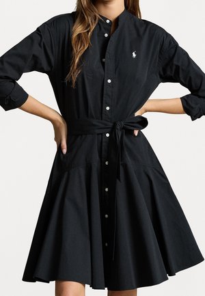 Mujer con un vestido camisa negro abotonado, con cinturón en la cintura y mangas largas remangadas, con un pequeño logo blanco en el pecho.