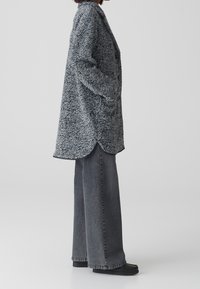 Abrigo gris de mezcla de lana con un patrón texturizado, diseño de doble botonadura y dos bolsillos laterales, combinado con jeans gris de pierna ancha y zapatos oscuros.