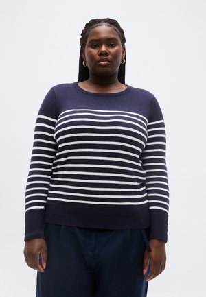 Vrouw met gevlochten haar, gouden oorringen en een marineblauwe trui met witte horizontale strepen, staand tegen een effen achtergrond.