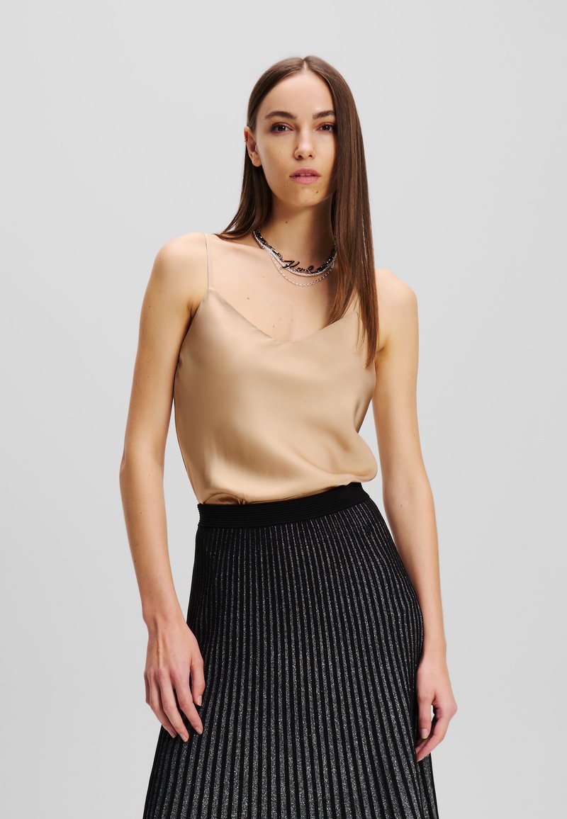 Beige satijnen top met dunne bandjes, gecombineerd met een zwarte geplisseerde rok met zilveren accenten. Model staat tegen een neutrale achtergrond.