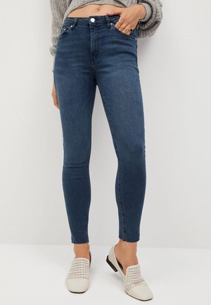 Mujer con vaqueros pitillo azul oscuro con dobladillos sin rematar, jersey gris corto y zapatos blancos de tejido trenzado sin cordones, de pie frente a un fondo neutro.