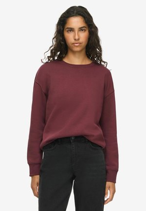 Femme aux longs cheveux bouclés portant un sweat-shirt bordeaux uni et un jean noir, debout devant un fond blanc.