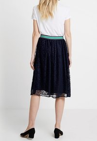 Granatowa koronkowa spódnica midi z czarno-zielonym paskiem, posiadająca zwiewny krój i fakturę, zestawiona z białą bluzką i czarnymi szpilkami.