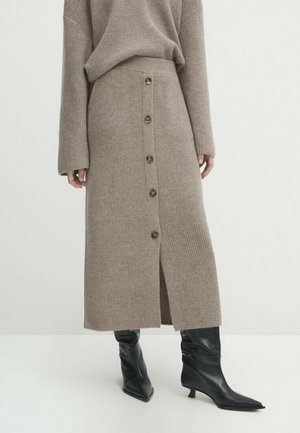 Jupe midi beige en maille avec boutons sur le devant, assortie à un pull coordonné et des bottes noires à talons moyens et bouts pointus, sur fond blanc.