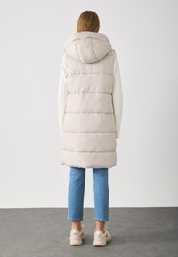 Lang beige dunvest med hette, med horisontal quilting, stylet med en kremfarget genser og blå jeans, komplett med beige sko.