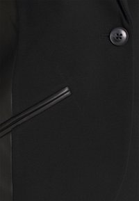 Détail d’un manteau en tissu noir avec une poche diagonale bordée de cuir et un grand bouton noir à quatre trous près du coin supérieur droit.