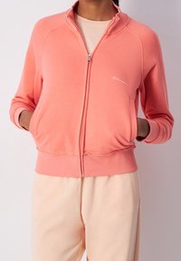 Sweat à zip couleur corail clair en tissu doux, avec des poignets et un ourlet côtelés, des poches avant et un petit logo blanc sur la poitrine.