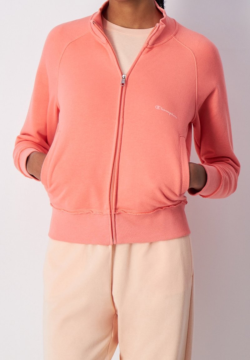 Sweat à zip couleur corail clair en tissu doux, avec des poignets et un ourlet côtelés, des poches avant et un petit logo blanc sur la poitrine.