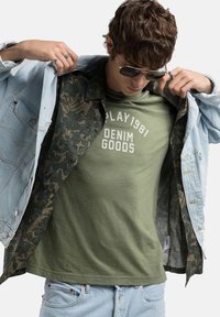 Chaqueta de denim clara con forro floral texturizado sobre una camiseta verde con letras blancas. Corte casual y relajado con costuras visibles.