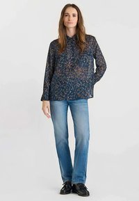 Flowy dark blue shirt with a vibrant abstract pattern, paired with light blue jeans and black shoes. Features button-down collar and two chest pockets.