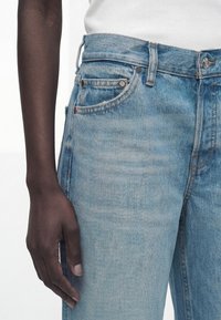 Άτομο με σκουρόχρωμο δέρμα που φοράει ανοιχτό μπλε τζιν και λευκό ριμπ τοπ, με το χέρι χαλαρό δίπλα στο σώμα σε λευκό φόντο.