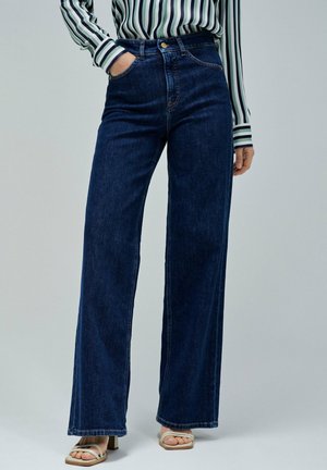 Jeans en denim à jambe large de couleur bleu foncé avec taille haute et poches avant, associés à une chemise rayée à manches longues et des sandales à talons neutres.
